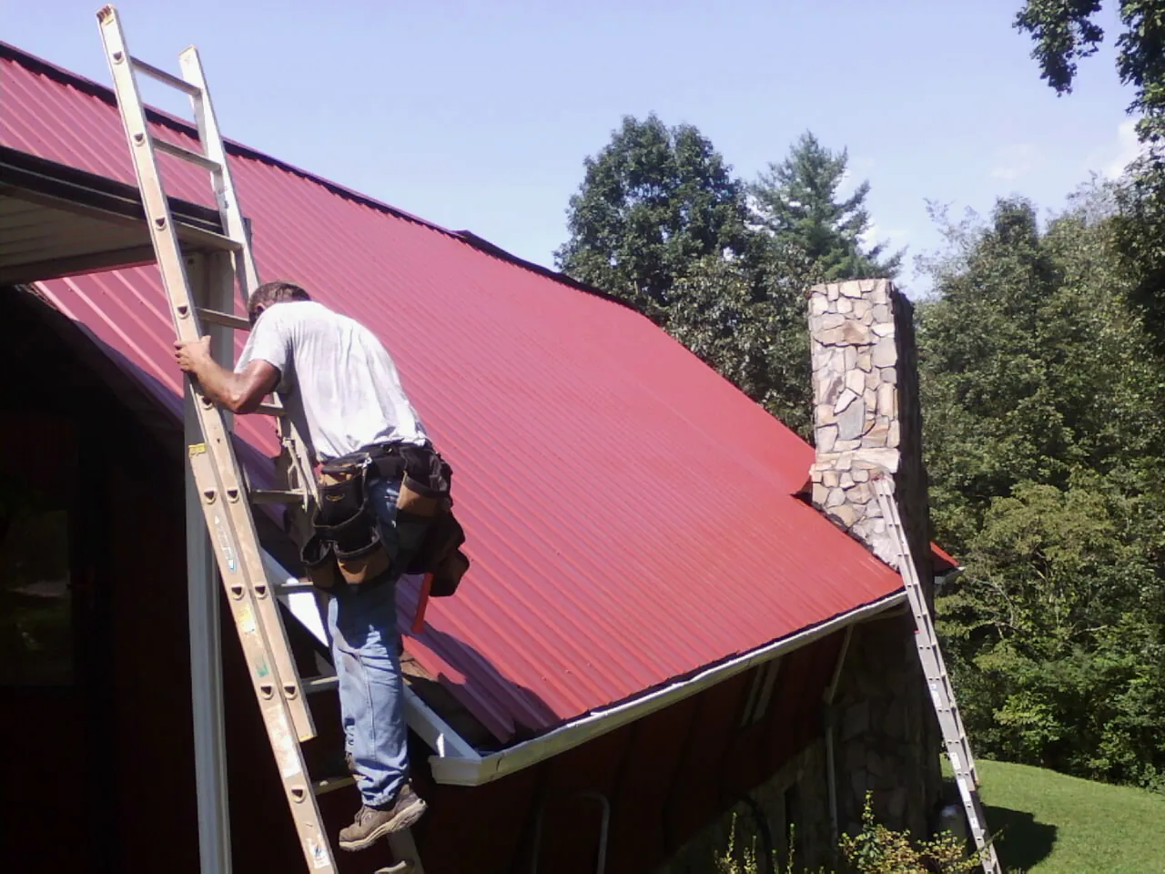 Roofer on a ladder beside a red metal roof and stone chimney — Soffit & Fascia Repair by Roofing Brothers in Steptoe